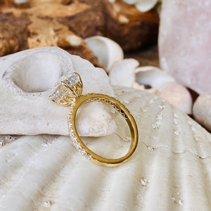 Pode incluir: Um anel de noivado de ouro com uma grande pedra central oval transparente e uma banda de diamantes menores.