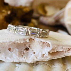 May include: A silver band ring with five emerald-cut diamonds set in a row. The ring is resting on a white, textured surface.