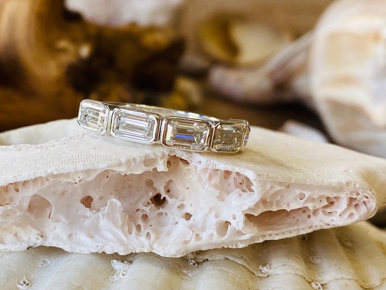 May include: A silver band ring with five emerald-cut diamonds set in a row. The ring is resting on a white, textured surface.