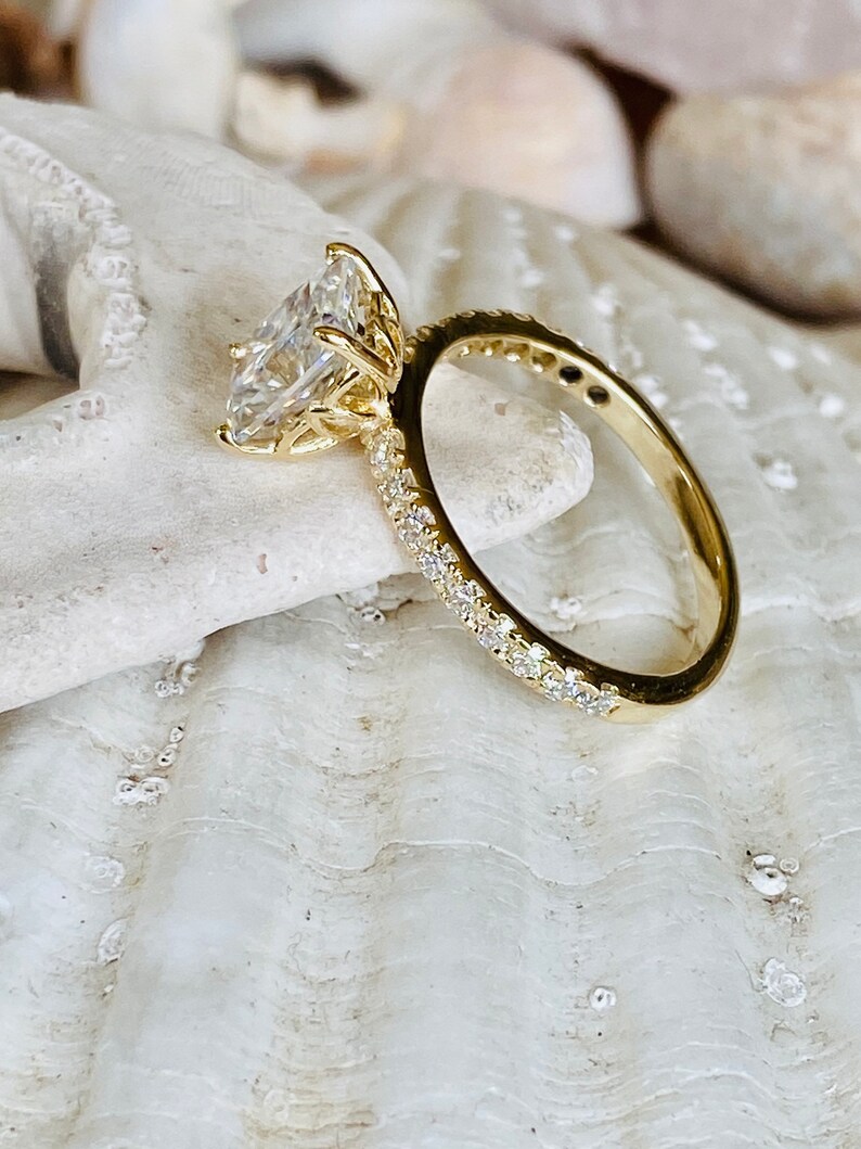 Pode incluir: Um anel de noivado de ouro com um grande diamante claro de forma quadrada. O anel tem uma banda de pequenos diamantes.