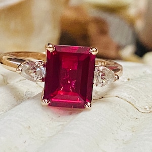 May include: A gold ring with a large, emerald-cut ruby gemstone and two pear-shaped diamonds on either side.