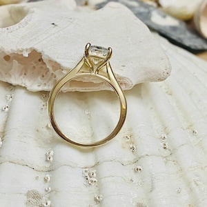May include: A gold engagement ring with a clear, round centre stone. The ring is set on a white seashell.