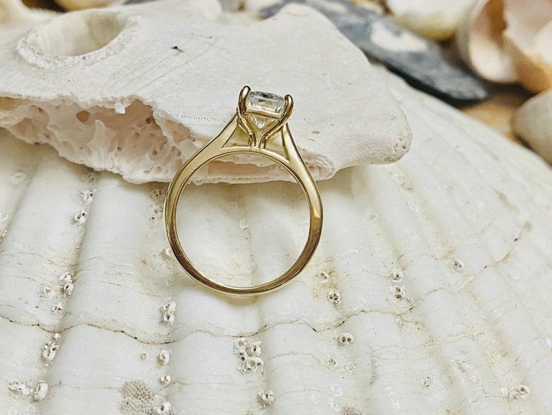May include: A gold engagement ring with a clear, round centre stone. The ring is set on a white seashell.