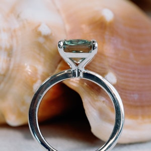 May include: A silver ring with a green gemstone set in a square shape. The ring is sitting on a seashell.