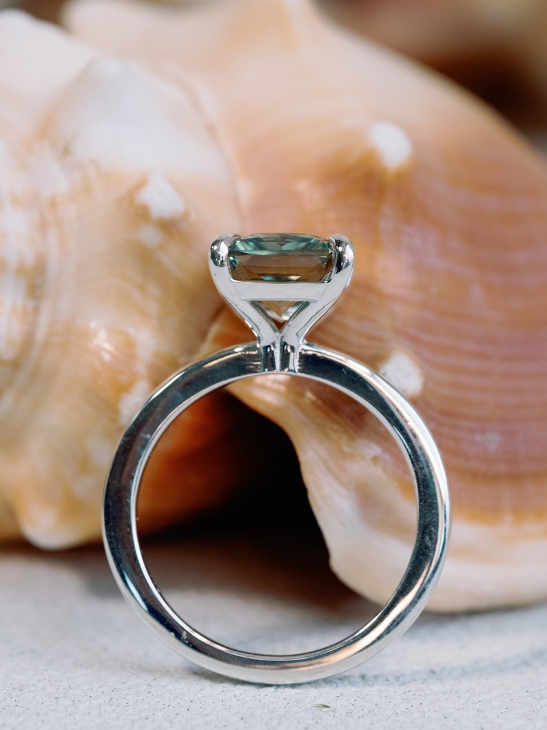 May include: A silver ring with a green gemstone set in a square shape. The ring is sitting on a seashell.