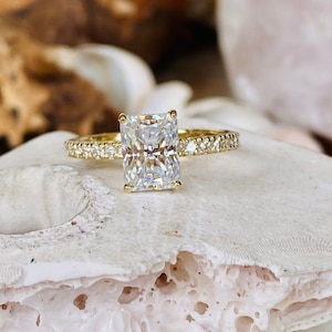 Pode incluir: Um anel de noivado de ouro com um grande diamante claro de forma quadrada e uma banda de diamantes menores.