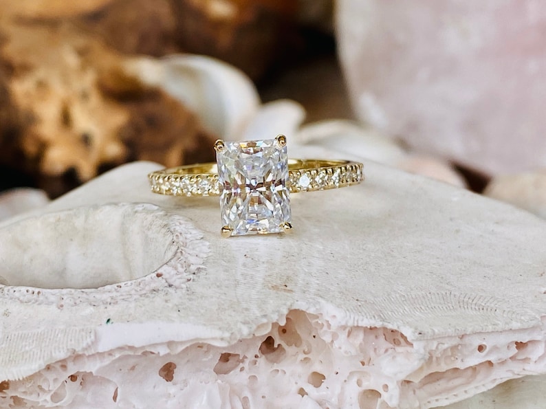 Pode incluir: Um anel de noivado de ouro com um grande diamante claro de forma quadrada e uma banda de diamantes menores.