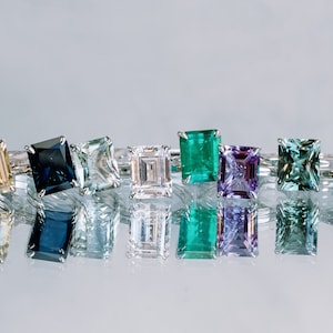 May include: A row of eight rings featuring various coloured gemstones. The gemstones are set in silver bands and all have an emerald cut. The colours of the gemstones are: yellow, blue, green, white, green, purple, green, and grey.