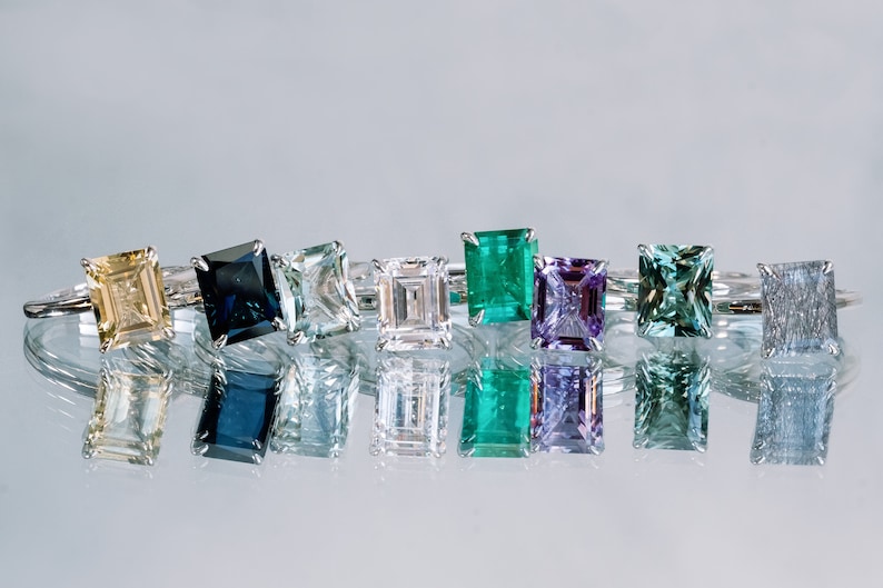 May include: A row of eight rings featuring various coloured gemstones. The gemstones are set in silver bands and all have an emerald cut. The colours of the gemstones are: yellow, blue, green, white, green, purple, green, and grey.