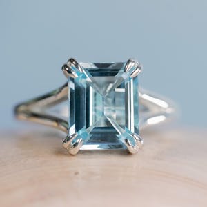 Puede incluir: Un anillo de plata con una gran gema rectangular de color azul claro. La gema está engastada con cuatro garras y la banda se divide en dos delicados hilos a cada lado. El anillo se muestra sobre una superficie clara.