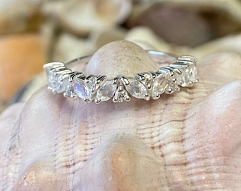 May include: A silver ring with a row of clear, marquise-cut gemstones and small round accent stones. The ring is set on a light-colored, textured surface, possibly a seashell. The gemstones are arranged in a repeating pattern.
