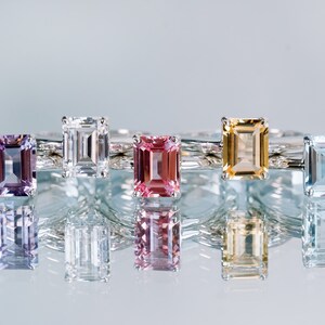 May include: Five emerald cut gemstone rings on a white background. The rings are set in silver and feature a variety of gemstones, including amethyst, white sapphire, pink sapphire, citrine, and aquamarine.