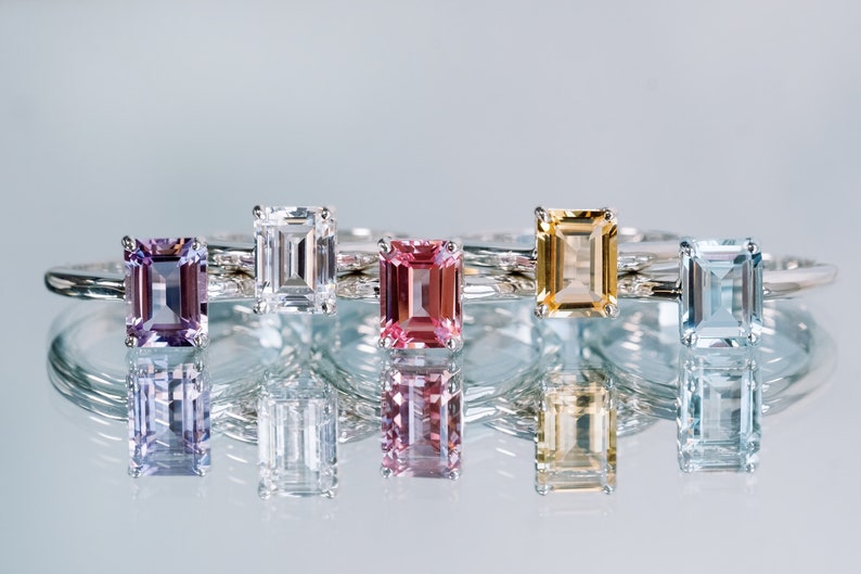 May include: Five emerald cut gemstone rings on a white background. The rings are set in silver and feature a variety of gemstones, including amethyst, white sapphire, pink sapphire, citrine, and aquamarine.