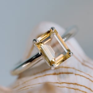 May include: A gold ring with a clear, rectangular, yellow-toned gemstone set in a silver band. The ring is resting on a white seashell.