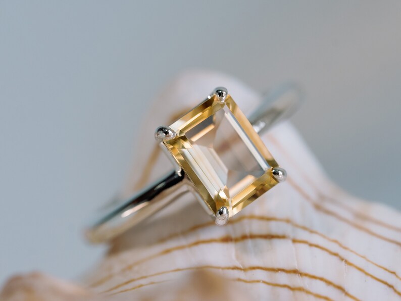 May include: A gold ring with a clear, rectangular, yellow-toned gemstone set in a silver band. The ring is resting on a white seashell.