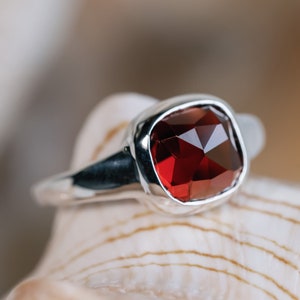 May include: A silver ring with a large, faceted, square-cut, deep red gemstone. The ring has a smooth, polished band, and the gemstone is set in a bezel setting. The ring is displayed on a seashell.