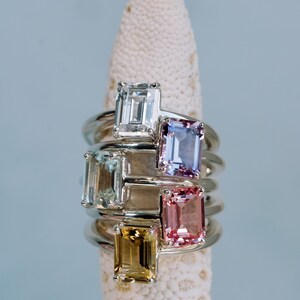 May include: Five rings with different colored gemstones, all set in silver. The gemstones are emerald cut and include clear, light blue, purple, pink, and yellow.