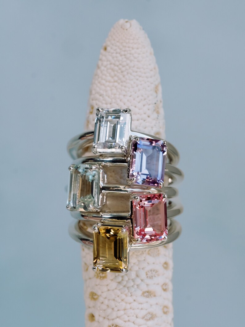 May include: Five rings with different colored gemstones, all set in silver. The gemstones are emerald cut and include clear, light blue, purple, pink, and yellow.