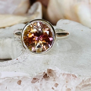 May include: A silver ring with a round, faceted gemstone. The gemstone is a mix of pink, orange, and yellow.