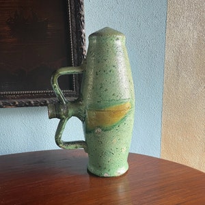 May include: A green ceramic pitcher with a unique design and a yellow stripe. The pitcher has a handle on the side and a spout on the top.