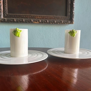 Può includere: Due portacandele in ceramica bianca con una figurina di rana verde. Ogni portacandele poggia su un piatto bianco con cerchi concentrici. I portacandele sono su una superficie di legno marrone scuro.