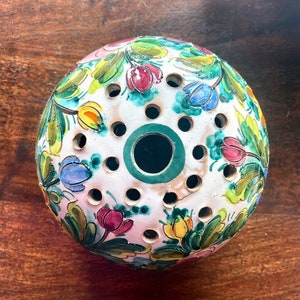 Può includere: Una sfera di ceramica bianca con un bordo verde e disegni floreali in tonalità di rosa, giallo, blu e verde. La sfera ha diversi fori attorno al centro.