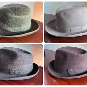 Può includere: Una collezione di quattro cappelli fedora in verde oliva, grigio chiaro, grigio antracite scuro e marrone scuro. Ogni cappello presenta una corona strutturata con piega centrale, una tesa corta e una fascia in tessuto abbinata alla base.