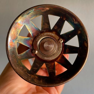 May include: A vintage, brass-colored, round, metal bottle stopper with a starburst design and a central gear mechanism. The stopper has a rainbow iridescent finish.