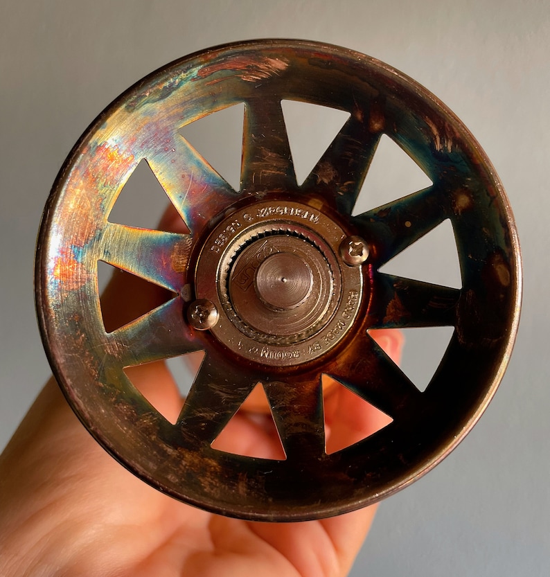 May include: A vintage, brass-colored, round, metal bottle stopper with a starburst design and a central gear mechanism. The stopper has a rainbow iridescent finish.