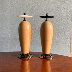 May include: A pair of wooden salt and pepper shakers with a unique design. The shakers are made of light wood with a natural grain pattern. The tops of the shakers are black and white and have a small metal knob. The bases of the shakers are made of metal with a gold finish.