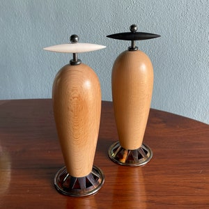 May include: Two wooden salt and pepper shakers with black and white tops. The shakers are made of light wood with a natural grain pattern. The bases are metal and have a decorative design.