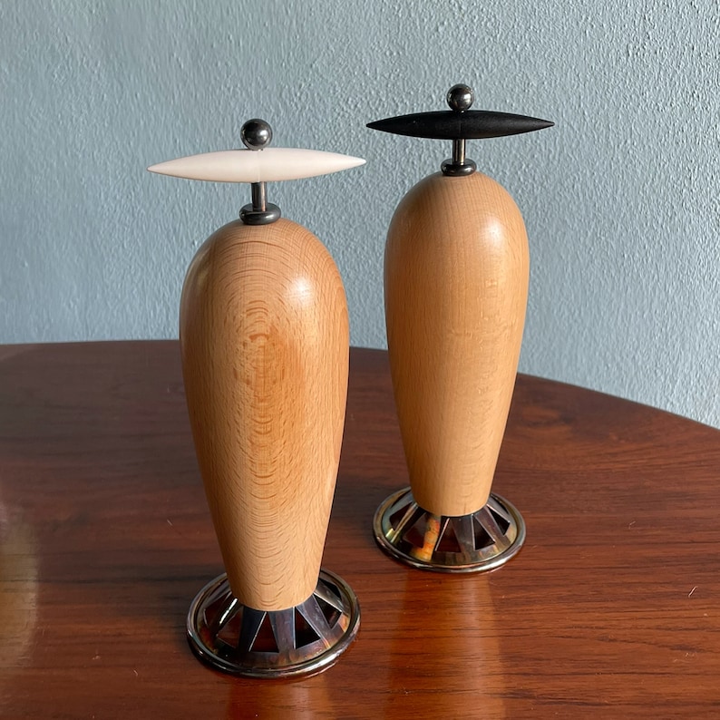 May include: Two wooden salt and pepper shakers with black and white tops. The shakers are made of light wood with a natural grain pattern. The bases are metal and have a decorative design.