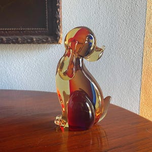 May include: A glass figurine of a dog in a sitting position. The dog is made of clear glass with shades of yellow, orange, and red. The figurine is about 6 inches tall.