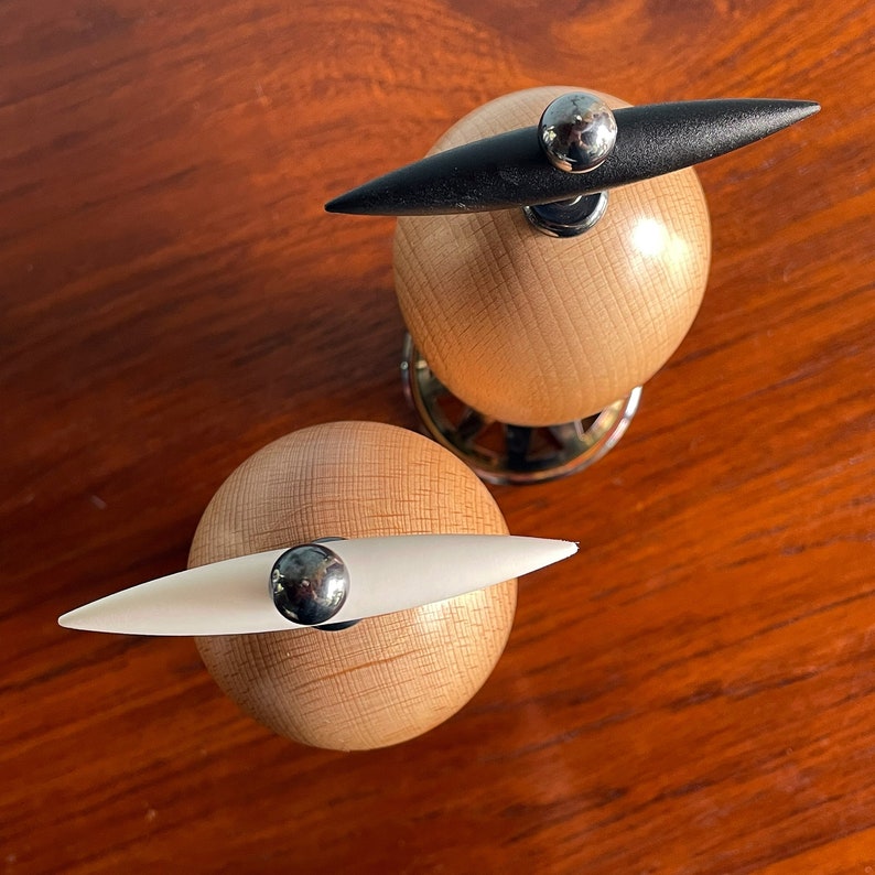 May include: Two wooden salt and pepper shakers with black and white handles. The shakers are round and have a wood grain pattern. The handles are attached to the top of the shakers and are shaped like a teardrop.