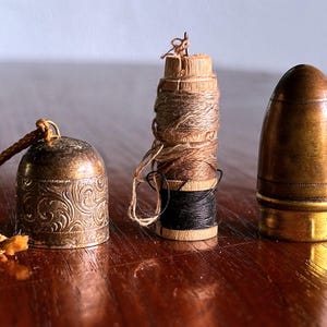 May include: Three antique sewing notions on a wooden surface. A bell-shaped thimble holder with a cord, a spool of thread with black and brown thread, and a bullet-shaped needle case. The items are made of brass and wood.
