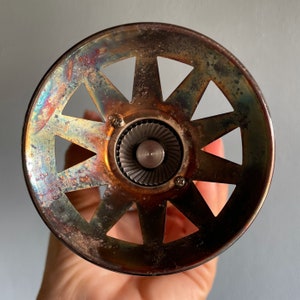 May include: A silver-toned metal bottle stopper with a starburst design and a central circular opening. The stopper has a worn, aged appearance with a rainbow-like iridescence.