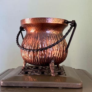 Könnte beinhalten: Ein gehämmerter Kupferkessel mit einem gedrehten Eisenhenkel und drei dekorativen Füßen. Der Kessel steht auf einer braunen Oberfläche.