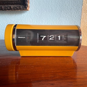 May include: A yellow digital clock with a black face and flip-up numbers. The clock reads 7:21 and has the word "Sankyo" printed on the side.