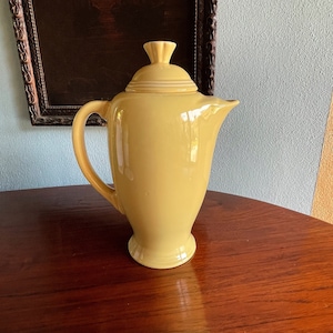 Può includere: Una caffettiera in ceramica gialla con un coperchio e un manico. La caffettiera è appoggiata su un tavolo di legno.