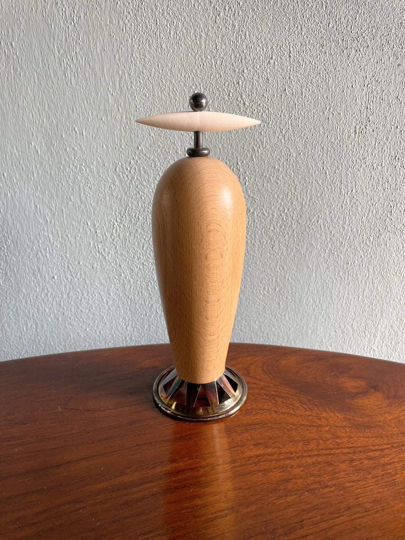 May include: A wooden bottle stopper with a white ceramic top and a metal base. The stopper is shaped like a vase and has a smooth, polished finish.