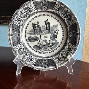 Puede incluir: Un plato antiguo decorativo con un borde floral en blanco y negro y una ilustración central. La ilustración representa varias figuras y las palabras "1 BIEN" y "D'UN". El plato se exhibe en un soporte de plástico transparente.
