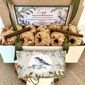 May include: A bird seed cake gift set in a white box with a green ribbon. The box contains several bird seed cakes with nuts and seeds, and a decorative card with a blue bird illustration. The box has the text "Enjoy!" and "Enjoy your bird seed cake gift set!"