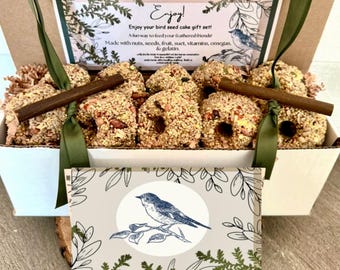 Puede incluir: Un juego de regalo de pastel de semillas para pájaros en una caja blanca con una cinta verde. La caja contiene varios pasteles de semillas para pájaros con nueces y semillas, y una tarjeta decorativa con una ilustración de pájaro azul.