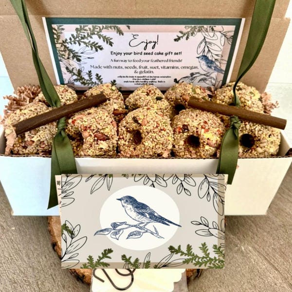 May include: A bird seed cake gift set in a white box with a green ribbon. The box contains several bird seed cakes with nuts and seeds, and a decorative card with a blue bird illustration. The box has the text "Enjoy!" and "Enjoy your bird seed cake gift set!"