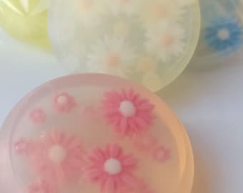 May include: Round, floral-themed soaps in shades of pink, yellow, and blue. Each soap features embedded flower designs, with the front-most soap showcasing pink flowers. The soaps are individually wrapped in clear plastic.