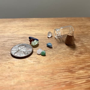 May include: A small glass bottle with a cork stopper, containing a variety of small, colorful stones. The bottle is lying on its side, and the stones have spilled out onto a wooden surface. A US penny is also visible in the image.
