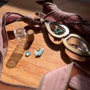 May include: A heart-shaped locket necklace with a glass window, revealing small green and blue stones. The necklace is made of brass and has a pink ribbon attached to it. There are also small stones and a cork on a wooden surface.