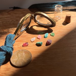 Puede incluir: Una pequeña lupa de bisagra de latón con una lente de vidrio transparente. La lupa está abierta y revela una colección de pequeñas piedras preciosas de colores. Las piedras preciosas son rosas, verdes y blancas. La lupa está sobre una superficie de madera.