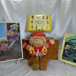 May include: A Cabbage Patch Kids doll dressed in a red and yellow cowboy outfit with a red hat. The doll is sitting on a wooden box with a book titled "Cabbage Patch Kids: A Cabbage Patch Kids Adventure" and a puzzle featuring Cabbage Patch Kids.