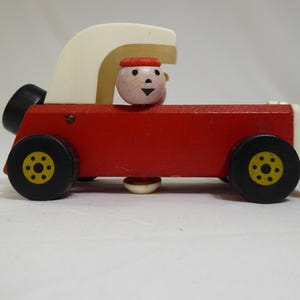 May include: A vintage wooden toy car with a red body, black wheels with yellow accents, and a white top. A small wooden figure with a red hat sits in the driver's seat. The toy is designed with simple, classic lines.